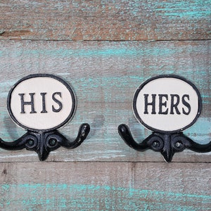 May include: Two black metal coat hooks with the words "HIS" and "HERS" in black lettering on a white oval background. The hooks are mounted on a weathered, rustic wooden surface with shades of blue and brown.