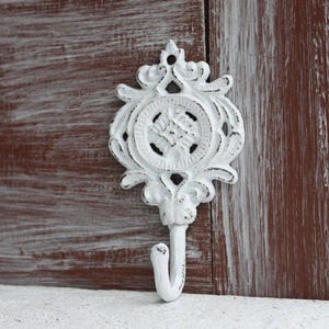May include: White cast iron hook with an ornate design. The hook has a floral pattern and a single hook at the bottom.