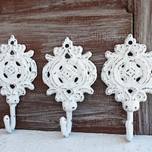 May include: Three white cast iron hooks with ornate designs. Each hook has a decorative scrollwork pattern and a single hook at the bottom.