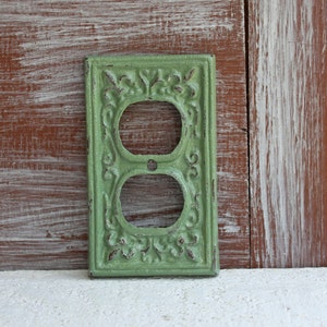 May include: A green metal outlet cover with an ornate design. The cover has a distressed finish and is designed to fit a standard double outlet.