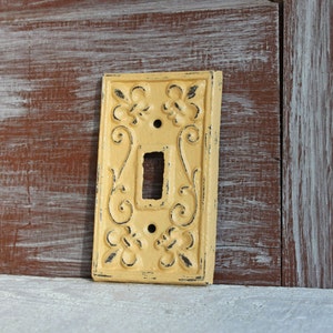 May include: A yellow cast iron light switch cover with an ornate design. The switch cover has a fleur-de-lis design and is distressed.