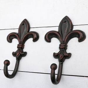 May include: Two black metal hooks with a fleur-de-lis design. The hooks are mounted on a white wooden surface.