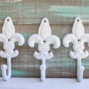 May include: Three white metal hooks shaped like fleur-de-lis, mounted on a wooden wall.