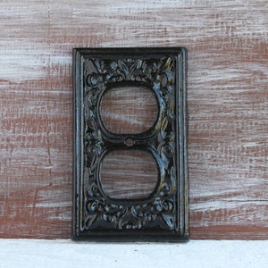 May include: Black metal outlet cover with an ornate design. The cover has a double outlet.