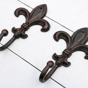May include: Two black cast iron hooks with a fleur-de-lis design. The hooks are mounted on a white wooden surface.