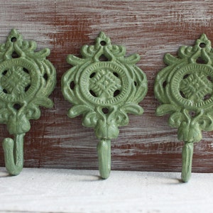 May include: Three green metal hooks with ornate designs. Each hook has a circular center with a decorative pattern and a curved hook extending downwards.