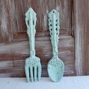 May include: A set of two decorative wall-mounted kitchen utensils, a fork and a spoon, in a light blue color with a distressed finish.