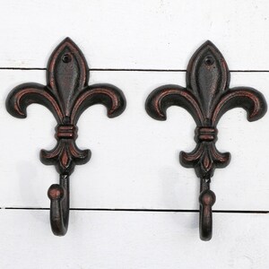 May include: Two black metal hooks with a fleur-de-lis design. The hooks are mounted on a white wooden surface.