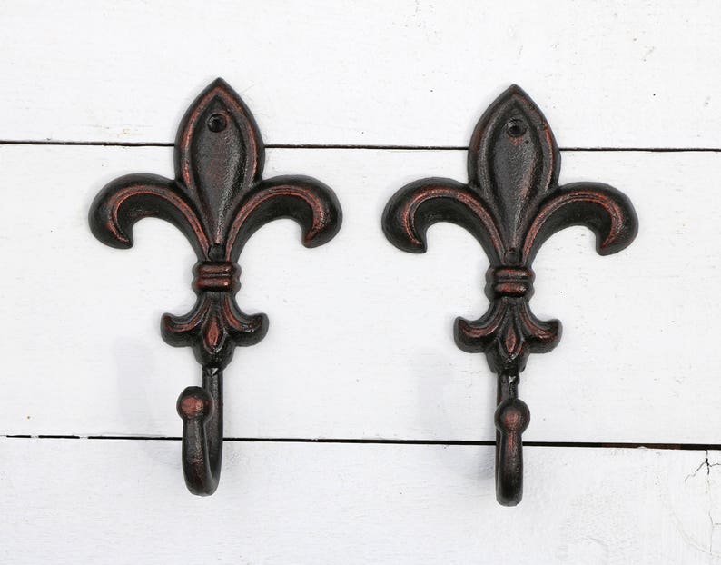 May include: Two black metal hooks with a fleur-de-lis design. The hooks are mounted on a white wooden surface.