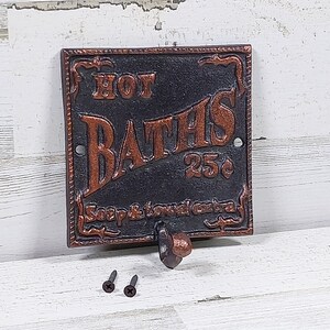 May include: A cast iron sign with the words "HOT BATHS 25¢" in copper-colored lettering. The sign has a hook below and includes two screws. The sign is square and has an ornate border.