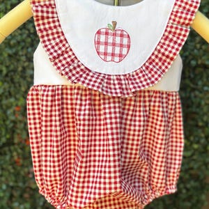 May include: A red and white gingham baby romper with a white collar featuring a red apple embroidery. The romper has a ruffled collar and a gathered bottom, perfect for a baby or toddler.