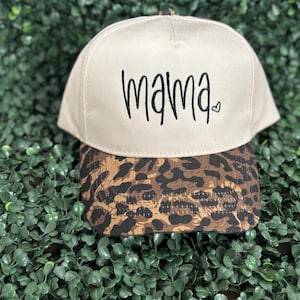 May include: Tan baseball cap with the word "mama" in black script and a small heart. The brim features a leopard print design. The cap is set against a backdrop of green foliage.
