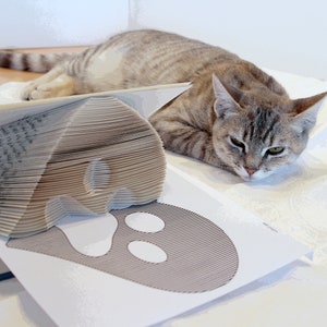 May include: A gray tabby cat is lying down next to a book that has been folded into the shape of a ghost. The ghost is outlined in black and has two white eyes.