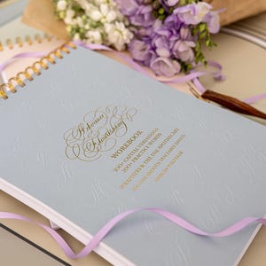 Op de afbeelding: Een lichtblauw werkboek met gouden letters en een bloemmotief, spiraalgebonden met gouden spiralen. Het werkboek ligt naast een boeket paarse en witte bloemen verpakt in bruin papier. Een paars lint is over het werkboek gedrapeerd.