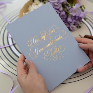 May include: A light blue rectangular notebook featuring gold foil calligraphy on the front cover that reads, Calligrapher You must make Haste. The notebook has a smooth matte finish and is held in a vertical orientation against a neutral background.