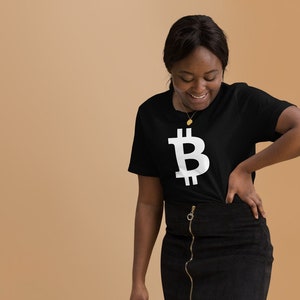 Könnte beinhalten: Eine Frau trägt ein schwarzes T-Shirt mit einem weißen Bitcoin-Symbol und einen schwarzen Jeansrock. Sie hält eine schwarze Handtasche mit einer goldenen Kette.
