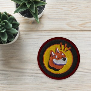 May include: A round embroidered patch with a black border and a red outer ring. The patch features a yellow background with a cartoon fox wearing a crown and the text "Woof!" in purple.