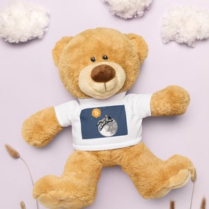 May include: A brown teddy bear wearing a white t-shirt with a graphic of a person floating in space with a Bitcoin symbol. The bear is lying on a light purple background with white clouds.
