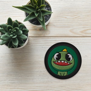 May include: A green and black embroidered patch featuring a cartoon dinosaur with big eyes and a toothy grin. The dinosaur is wearing a yellow hat.