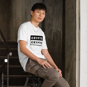 Puede incluir: Una persona lleva una camiseta blanca con el texto "CRYPTO" impreso en negro sobre un rectángulo negro. La persona está sentada en un taburete negro y lleva pantalones cargo marrones.