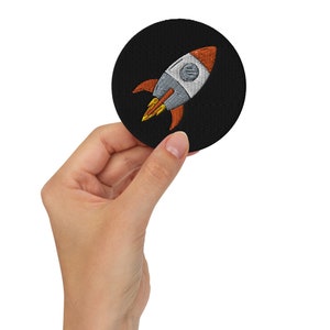 May include: A black embroidered patch featuring a white and orange rocket ship design.