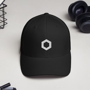 May include: Black baseball cap with a white hexagonal logo on the front. The cap is made of a soft material and has a curved brim. Headphones and a notebook are in the background.