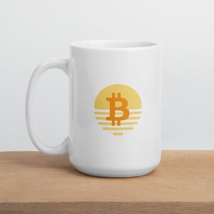 Pode incluir: Caneca de cerâmica branca com um gráfico de um sol amarelo com um símbolo Bitcoin laranja dentro. O sol tem listras laranja por baixo.