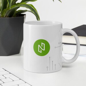 Puede incluir: Taza de cerámica blanca con un círculo verde que contiene la letra "N" y un diseño de circuito gris.