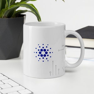 May include: White ceramic mug with a blue and gray design featuring the logo of the cryptocurrency Cardano. The logo is a circle with a central dot and concentric circles of dots around it.