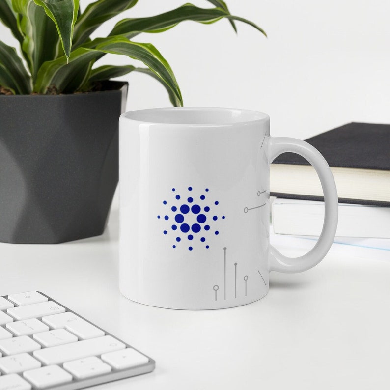 May include: White ceramic mug with a blue and gray design featuring the logo of the cryptocurrency Cardano. The logo is a circle with a central dot and concentric circles of dots around it.
