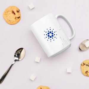 May include: White ceramic mug with a blue circle logo and a small key symbol on the side. The mug is surrounded by sugar cubes and cookies on a white surface.