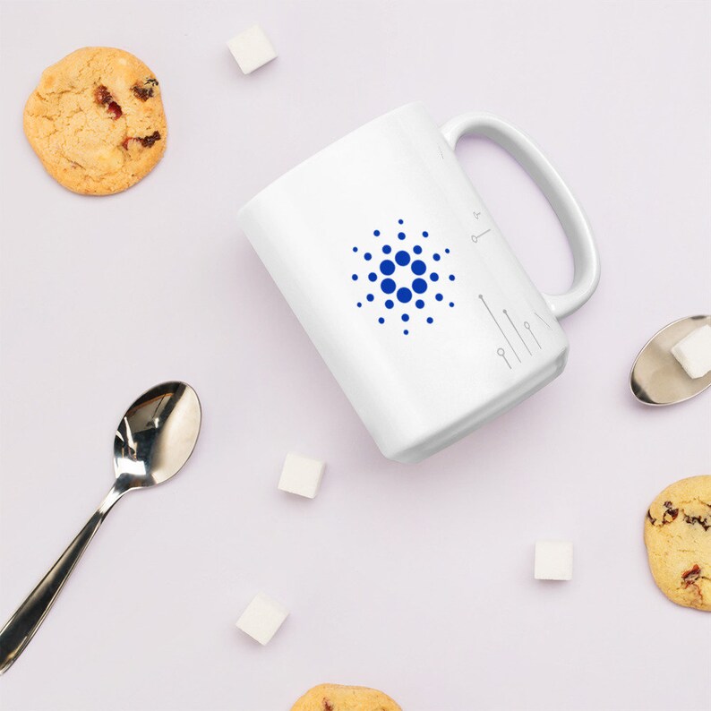 May include: White ceramic mug with a blue circle logo and a small key symbol on the side. The mug is surrounded by sugar cubes and cookies on a white surface.