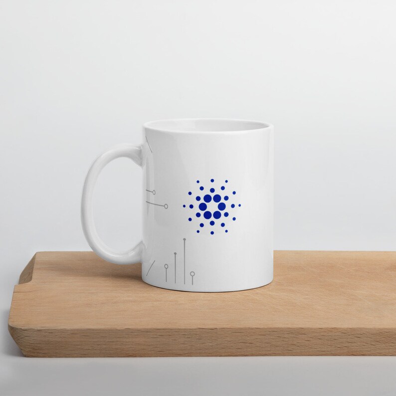 May include: White ceramic mug with a blue and white logo of a circle with dots around it. The logo is on the right side of the mug. The mug is on a wooden surface.