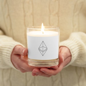 May include: A burning candle in a clear glass jar. The candle features a white label with a black Ethereum logo. The candle is held in both hands, with a cream-coloured sweater in the background.