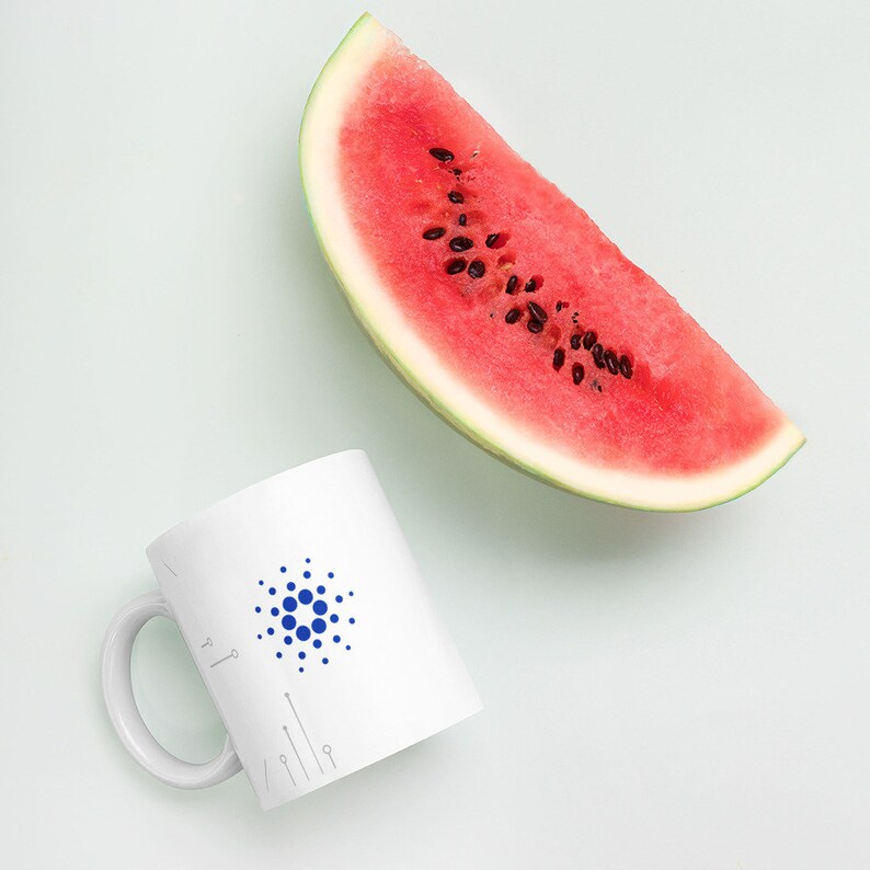 May include: A white ceramic mug with a blue and white logo of a circle with dots. The mug is next to a slice of watermelon on a white background.