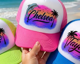 May include: Three trucker hats with white front panels and colored mesh backs in neon green, hot pink, and bright blue. Each hat features a sunset graphic with palm trees and a name printed in black script: Sophie, Chelsea, and Taylor.