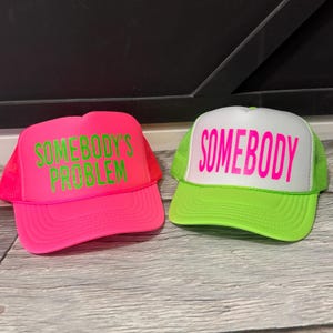 May include: Two trucker hats. One is bright pink with the text "SOMEBODY'S PROBLEM" in green. The other is white with the word "SOMEBODY" in pink, and a neon green brim and mesh back.