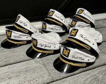 May include: A collection of white captain hats featuring black brims, gold braided cords, and embroidered gold anchor emblems. Each hat is personalized with black script text including Cpt Kyle, Cpt Eric, Coach Seth, Daddy Dara, Cpt Jared, Chief Emo, Cpt Lee, The Groom, and Best Man.