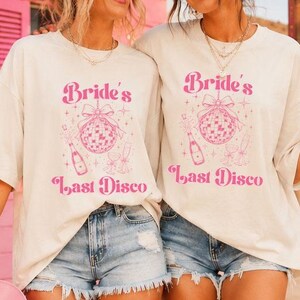 May include: An off-white short-sleeve t-shirt featuring a pink graphic print that includes the text "Bride's Last Disco," a disco ball with a bow, a champagne bottle, and two champagne flutes, all surrounded by small star-shaped accents.