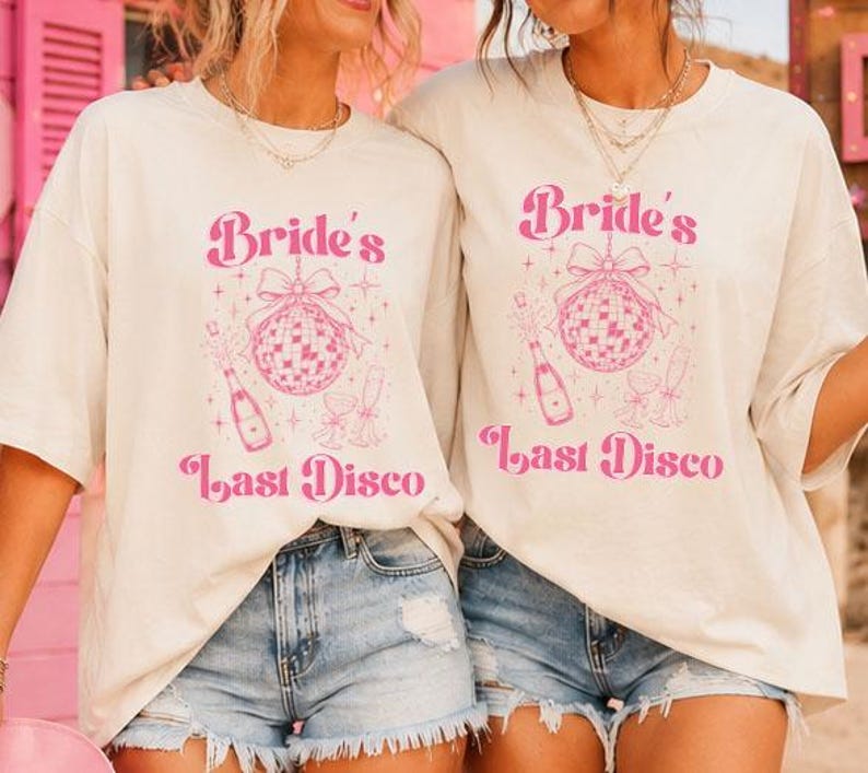 May include: An off-white short-sleeve t-shirt featuring a pink graphic print that includes the text "Bride's Last Disco," a disco ball with a bow, a champagne bottle, and two champagne flutes, all surrounded by small star-shaped accents.