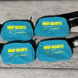 May include: Four teal fanny packs with black straps and zippers. Each pack has the text "NAUR REGRETS The Sundholms" in yellow. The packs are arranged on a gray wooden surface.