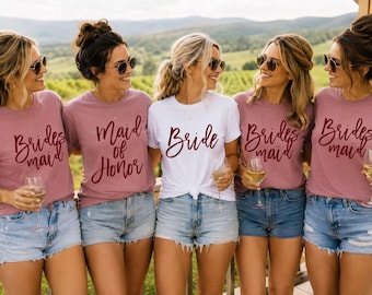 May include: Five t-shirts worn with denim shorts. Four shirts are dusty rose with dark red text reading Bridesmaid, and one center shirt is white with dark red text reading Bride. Each shirt features short sleeves and a crew neckline.