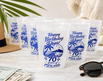 May include: A set of clear plastic cups featuring a blue graphic print of a tropical beach scene with palm trees, waves, and a mountain. The text reads Happy Birthday Pops! 70 years of pura vida. The cups are arranged in a group on a light surface.