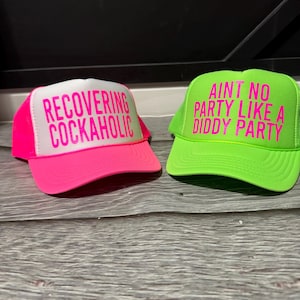 May include: Two trucker hats. One is bright pink with the text "RECOVERING COCKAHOLIC" in pink. The other is neon green with the text "AINT NO PARTY LIKE A DIDDY PARTY" in pink. Both hats have a white front panel.