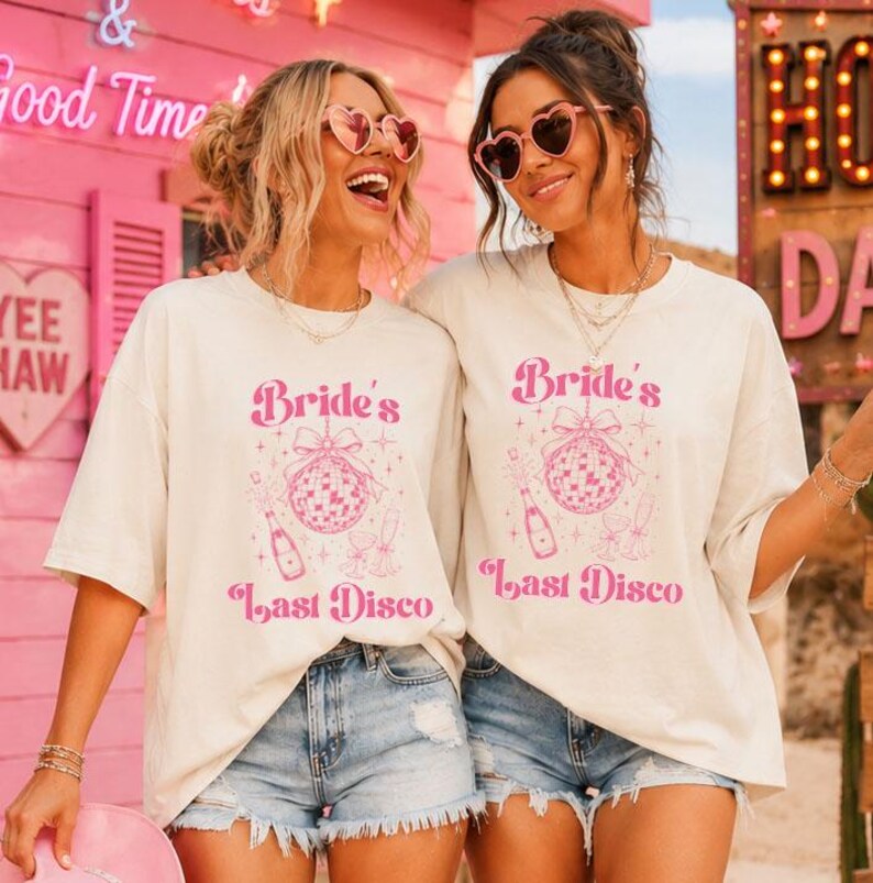 May include: Two off-white short-sleeve t-shirts featuring a pink graphic print of a disco ball with a bow, a champagne bottle, and the text "Bride's Last Disco". The shirts are paired with light blue denim shorts and heart-shaped sunglasses.