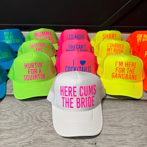 May include: A collection of trucker hats in various colors, including blue, green, pink, yellow, and orange. Each hat features bold, neon text with humorous phrases. The hats have a mesh back and a curved brim.
