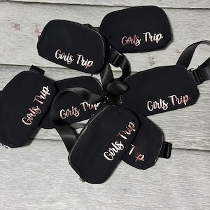 May include: Five black fanny packs with adjustable straps. Each pack has the text "Girls Trip" embroidered in rose gold.