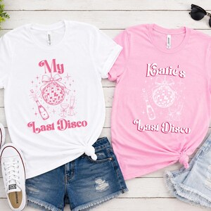 May include: A white t-shirt with pink text reading My Last Disco and a pink graphic of a disco ball and champagne, and a pink t-shirt with white text reading Katie's Last Disco and a white graphic of a disco ball and champagne.