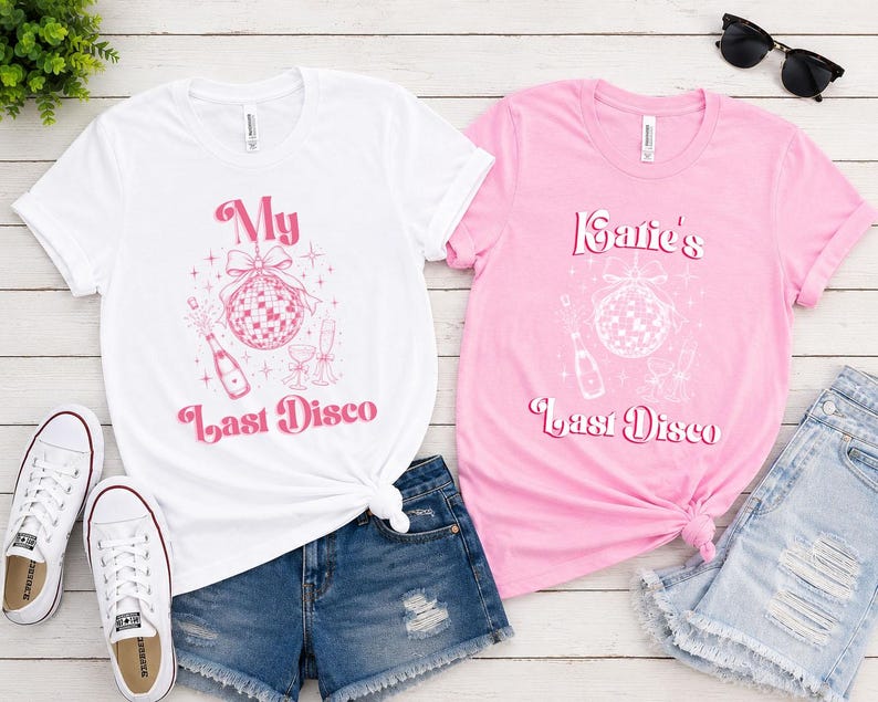 May include: A white t-shirt with pink text reading My Last Disco and a pink graphic of a disco ball and champagne, and a pink t-shirt with white text reading Katie's Last Disco and a white graphic of a disco ball and champagne.