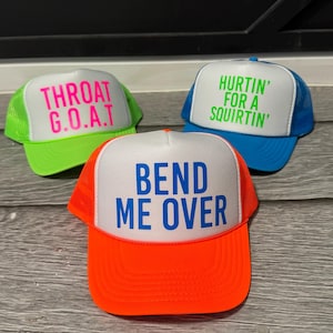 May include: Three trucker hats with white front panels and mesh backs. The hats have different colored brims and text: one with a neon green brim and the text "THROAT G.O.A.T", one with a blue brim and the text "HURTIN' FOR A SQUIRTIN'", and one with an orange brim and the text "BEND ME OVER".